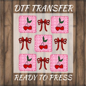 May include: DTF transfer sheet featuring a repeating pattern of red cherries and red bows on pink gingham squares. The text "DTF TRANSFER" and "READY TO PRESS" are printed in white.