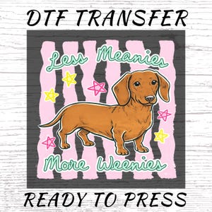 Peut inclure: Transfert DTF avec une illustration de chien teckel marron. Le motif comprend le texte "Less Meanies More Weenies" dans une police ludique, avec des rayures roses et blanches en arrière-plan, et des étoiles jaunes et roses. Les mots "READY TO PRESS" sont en bas.
