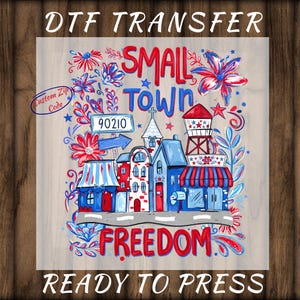 May include: DTF transfer featuring a colorful illustration of a small town with the words "Small Town Freedom" in red. The design includes buildings, flowers, and a water tower, with the text "Ready to Press."