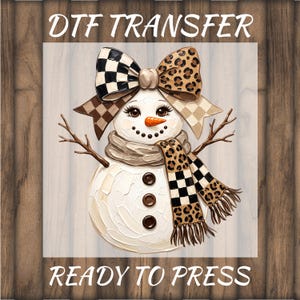 May include: A DTF TRANSFER READY TO PRESS graphic featuring a snowman with a large bow on its head, wearing a scarf with leopard print and black and white checkered patterns. The snowman has twig arms, three brown buttons, and an orange nose.
