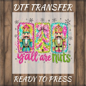 May include: A DTF transfer featuring two nutcracker figures and a central Christmas tree with a pink and white striped bow. The design includes snowflakes and the text Y'ALL ARE NUTS in pink and green. The graphic is ready to press.