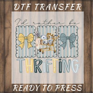 May include: DTF transfer design with the text "I'd rather be thrifting." The design features a shopping cart filled with vintage items, bows, and a striped background. The words "Ready to press" are also included.