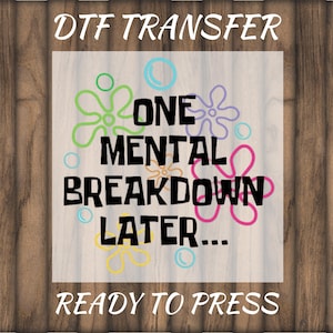 May include: DTF TRANSFER graphic with the text "ONE MENTAL BREAKDOWN LATER... READY TO PRESS" in black, surrounded by colorful flower and bubble designs. The background is a light wood grain.