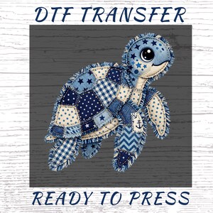 May include: A blue patchwork turtle design with stars, polka dots, and anchor details. The text "DTF TRANSFER" is at the top, and "READY TO PRESS" is at the bottom. The turtle is on a gray wooden background.