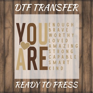May include: A DTF transfer design with the words "YOU ARE" in brown and tan, with a heart graphic. The words "ENOUGH, BRAVE, WORTHY, LOVED, AMAZING, STRONG, CAPABLE, SMART, KIND" are listed vertically. The text "READY TO PRESS" is at the bottom.