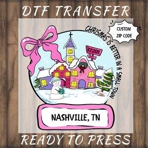 May include: DTF transfer with a snow globe design showing a winter town scene, a snowman, and a Christmas tree. Text includes "Christmas is better in a small town" and "Nashville, TN". Features a custom zip code option.