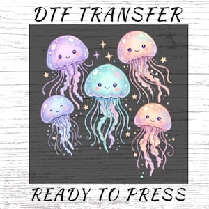 May include: A DTF transfer sheet featuring five cartoon jellyfish in pastel shades of purple, blue, and pink with smiling faces and long tentacles. The design includes small star accents on a dark background with the text DTF TRANSFER READY TO PRESS.