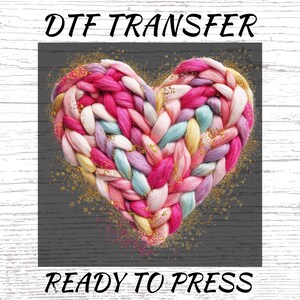 May include: A colorful heart-shaped design made of woven yarn in shades of pink, purple, blue, and yellow. The words "DTF TRANSFER" and "READY TO PRESS" are written in black script above and below the heart. Gold glitter accents the design.