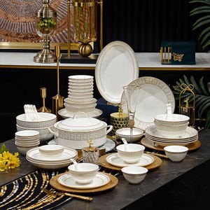 May include: A luxurious white and gold dinnerware set, including plates, bowls, and serving dishes. The set features a gold geometric pattern and trim, with a variety of sizes and shapes for a complete dining experience. The set is arranged on a dark surface.
