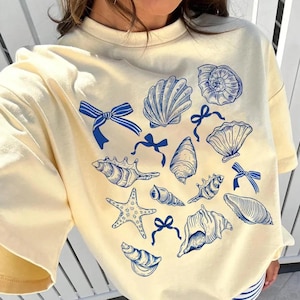 May include: Cream-colored t-shirt featuring a blue seashell and bow design. The graphic includes various seashell illustrations and bow accents. The shirt has short sleeves and a crew neck.