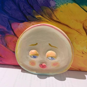 May include: A hand-painted, ceramic dish in the shape of a face with a sad expression. The dish is off-white with blue eyes, pink cheeks, and a red mouth. The background is a colorful abstract design.