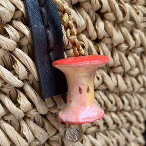 May include: A close-up of a straw bag with a decorative apple charm. The charm is painted with red, pink, and yellow hues, and has a small clear bead attached. The bag has a black leather strap.
