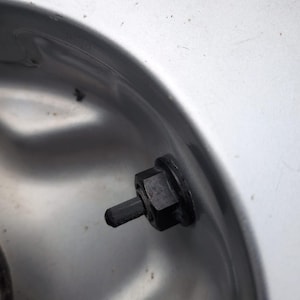 May include: A black rubber tire valve stem with a hexagonal base nut protruding from a smooth, metallic silver wheel rim. The valve stem is cylindrical with a textured tip, positioned centrally within the curved surface of the metal wheel component.