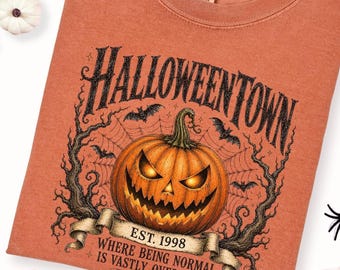 Może przedstawiać: T-shirt w kolorze wypalonej pomarańczy z grafiką przedstawiającą dynię, bezlistne drzewa i pajęczyny. Tekst głosi HALLOWEENTOWN EST. 1998 WHERE BEING NORMAL IS VASTLY OVERRATED. Koszulka ma okrągły dekolt oraz metkę marki Comfort Colors.