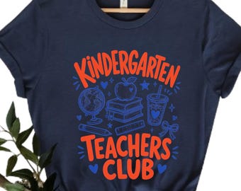 Może przedstawiać: Granatowa koszulka z krótkim rękawem z pomarańczowym napisem "KINDERGARTEN TEACHERS CLUB" oraz niebieskimi grafikami liniowymi przedstawiającymi globus, stos książek, jabłko, ołówek, linijkę, kubek, kokardę i małe serca z przodu.