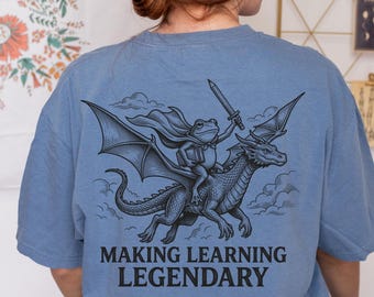 Może przedstawiać: Niebieski t-shirt z czarną grafiką na plecach przedstawiającą żabę w pelerynie, trzymającą miecz i książkę, jadącą na smoku. Pod ilustracją znajduje się napis "MAKING LEARNING LEGENDARY" wykonany pogrubioną, czarną czcionką bezszeryfową.