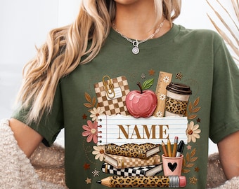 Może przedstawiać: Wojskowo-zielony t-shirt z nadrukiem graficznym przedstawiającym przybory szkolne, w tym czerwone jabłko, kubek kawy, stos książek, linijkę i ołówek. Środek wzoru zawiera grafikę papieru w linie z napisem NAME.