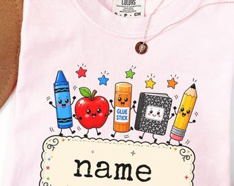 Może przedstawiać: Jasnoróżowy t-shirt z grafiką przedstawiającą niebieską kredkę, czerwone jabłko, pomarańczowy klej w sztyfcie, czarno-biały zeszyt oraz żółty ołówek z kolorowymi gwiazdkami. Pod postaciami znajduje się kremowa etykieta z napisem name.