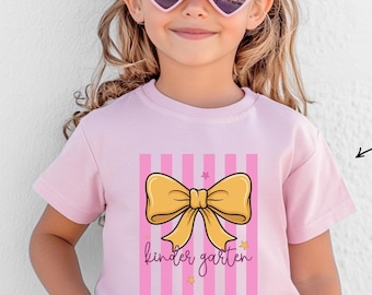 Może przedstawiać: Jasnoróżowy t-shirt z krótkim rękawem z grafiką dużej żółtej kokardy umieszczonej centralnie na pionowych różowych paskach. Napis "kinder garten" jest wydrukowany kursywą w kolorze czarnym pod kokardą, w towarzystwie małych żółtych gwiazdek.