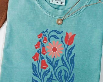 Może przedstawiać: Turkusowy t-shirt z krótkim rękawem i metką Comfort Colors przy szyi. Koszulka posiada nadruk graficzny przedstawiający czerwone kwiaty w kształcie dzwonków, różowy stokrotkę oraz czerwony tulipan z ciemnoniebieskimi liśćmi. Na tkaninie spoczywa złoty naszyjnik z małą okrągłą zawieszką.