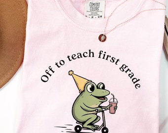 Może przedstawiać: Jasnoróżowy t-shirt z grafiką zielonej żaby w żółtej czapce imprezowej, jadącej na hulajnodze i trzymającej napój. Tekst nad żabą brzmi "Off to teach first grade". Koszulka ma okrągły dekolt i metkę Comfort Colors.
