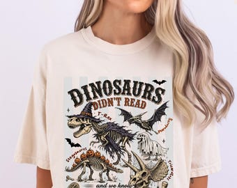 Może przedstawiać: Koszulka z krótkim rękawem w kolorze kości słoniowej z nadrukiem graficznym zawierającym tekst DINOSAURS DIDN'T READ oraz what happened to them. Wzór zawiera ilustracje szkieletów tyranozaura w kapeluszu czarownicy, pterodaktyla, stegozaura z dyniami oraz triceratopsa.