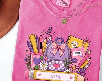 Może przedstawiać: Różowy t-shirt z nadrukiem graficznym przedstawiającym fioletowy plecak z kokardą, piórnik z przyborami szkolnymi, notatnik z napisem NOTES oraz duży żółty ołówek z tekstem NAME. Koszulka posiada metkę marki Comfort Colors.