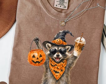 Może przedstawiać: Brązowy t-shirt z krótkim rękawem z grafiką szopa pracza w kapeluszu czarownicy, okularach przeciwsłonecznych w kształcie serc i pomarańczowej bandanie w nietoperze. Szop trzyma wiaderko w kształcie dyni i mrożoną kawę. Metka Comfort Colors.