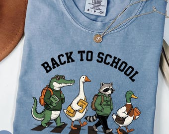 Może przedstawiać: Niebieski t-shirt z krótkim rękawem, okrągłym dekoltem i nadrukiem przedstawiającym krokodyla, gęś, szopa pracza i kaczkę idących po przejściu dla pieszych. Nad zwierzętami trzymającymi książki i plecaki widnieje napis BACK TO SCHOOL.