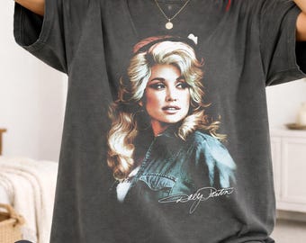 May include: A charcoal grey short-sleeve t-shirt featuring a vintage-style graphic print of Dolly Parton with blonde hair and a denim jacket. The shirt has a crew neckline and a relaxed fit, with the signature Dolly Parton printed in white script.