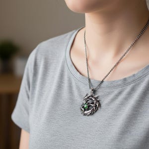 May include: A silver-colored pendant necklace featuring a dragon design. The dragon is wrapped around a green gemstone. The necklace is worn on a silver chain. The person is wearing a gray t-shirt.