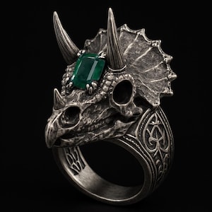 May include: A silver ring with a triceratops skull design, featuring a green emerald-cut gemstone. The ring's band is intricately carved with patterns. The design is detailed and the ring is a bold statement piece, suitable for a unique style.