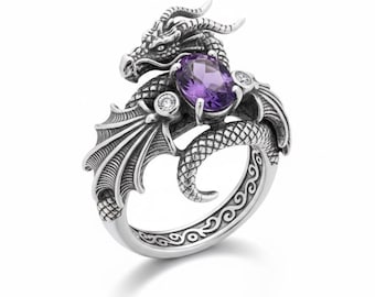 May include: A silver dragon ring with an oval purple gemstone and two smaller clear stones. The dragon has detailed wings and scales, with a decorative band. The ring is set against a white background.