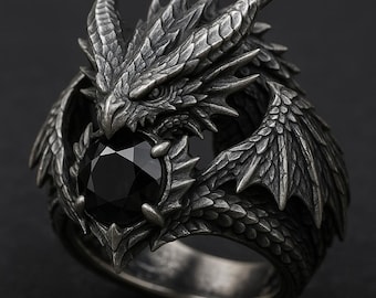 May include: A detailed silver dragon ring with a black gemstone. The dragon's head and wings are intricately sculpted, with the gemstone held in its jaws. The ring has a dark, antique finish.