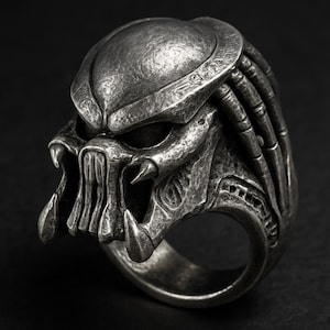 May include: A detailed, silver-toned ring featuring the sculpted face of a Predator. The ring showcases the creature's distinctive mandibles, dreadlocks, and helmet design. The ring is a bold statement piece, perfect for fans of the sci-fi film.
