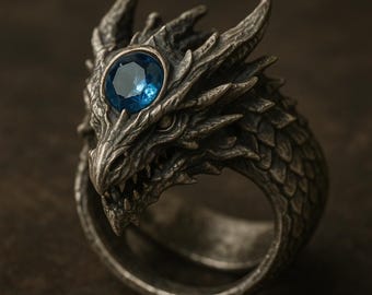 Può includere: Anello a forma di drago argentato con dettagli intricati. La testa del drago presenta un occhio di gemma blu. L'anello è progettato con un aspetto strutturato e squamoso, che mostra un tema fantasy. Un gioiello unico.