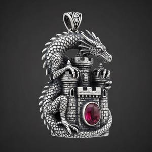 Puede incluir: Colgante plateado con un dragón sobre un castillo con una gema roja. El dragón tiene escamas detalladas y agarra las torres del castillo. El colgante tiene una fianza decorativa con una pequeña gema.
