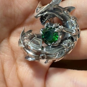 May include: A silver dragon pendant necklace with a detailed design. The dragon is wrapped around a large, round, emerald-green gemstone. The pendant is suspended from a delicate silver chain, creating a striking piece of jewelry.
