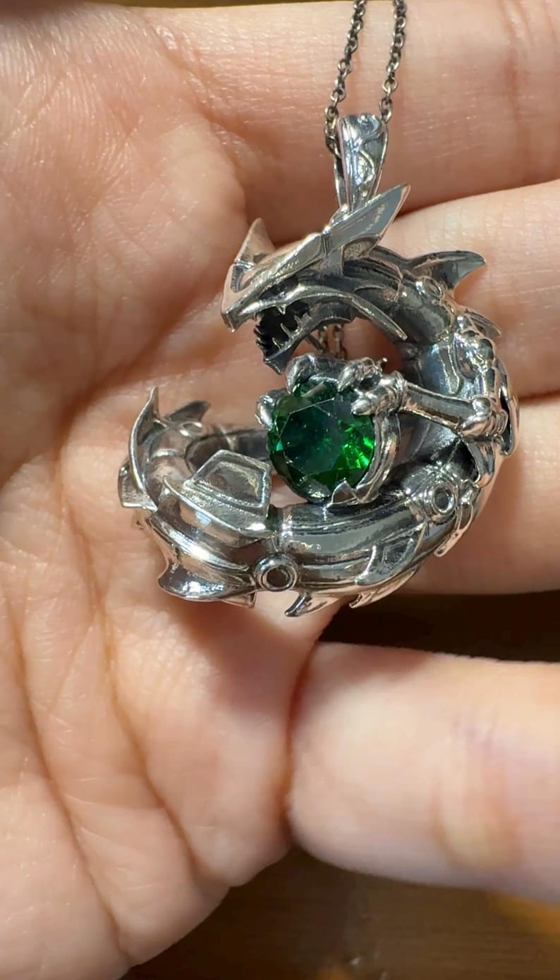 May include: A silver dragon pendant necklace with a detailed design. The dragon is wrapped around a large, round, emerald-green gemstone. The pendant is suspended from a delicate silver chain, creating a striking piece of jewelry.