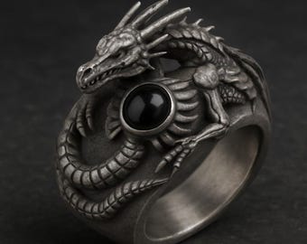 Può includere: Un anello argentato con un design dettagliato di un drago. Il drago avvolge l'anello, con la testa e la coda che si incontrano. Una pietra nera e rotonda è incastonata al centro. L'anello ha una finitura strutturata e antica.