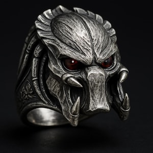 May include: A detailed silver-colored ring featuring a Predator alien design. The ring showcases the creature's distinctive face with sharp teeth and glowing red eyes. The ring is set against a dark background.