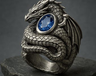 Può includere: Anello a forma di drago argentato con una grande gemma blu ovale. Il corpo del drago si avvolge attorno all'anello, con squame e ali dettagliate. La gemma è incastonata in una lunetta d'argento.