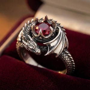 Puede incluir: Un anillo de dragón plateado con una gema roja grande y redonda. El anillo presenta un diseño detallado de dragón con alas y escamas. La gema está engastada en un engaste en forma de corona.