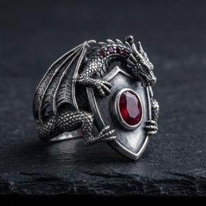 Könnte beinhalten: Silberfarbener Drachenring mit Schilddesign. Der Drache hat detaillierte Schuppen, Flügel und rote Edelsteinakzente. Das Schild hat einen großen, ovalen, roten Edelstein. Der Ring ist vor dunklem Hintergrund.