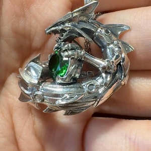 May include: A silver pendant necklace featuring a detailed, stylized shark design. The shark holds a large, round, emerald-green gemstone. The pendant hangs from a delicate silver chain, showcasing intricate metalwork and a unique, aquatic theme.
