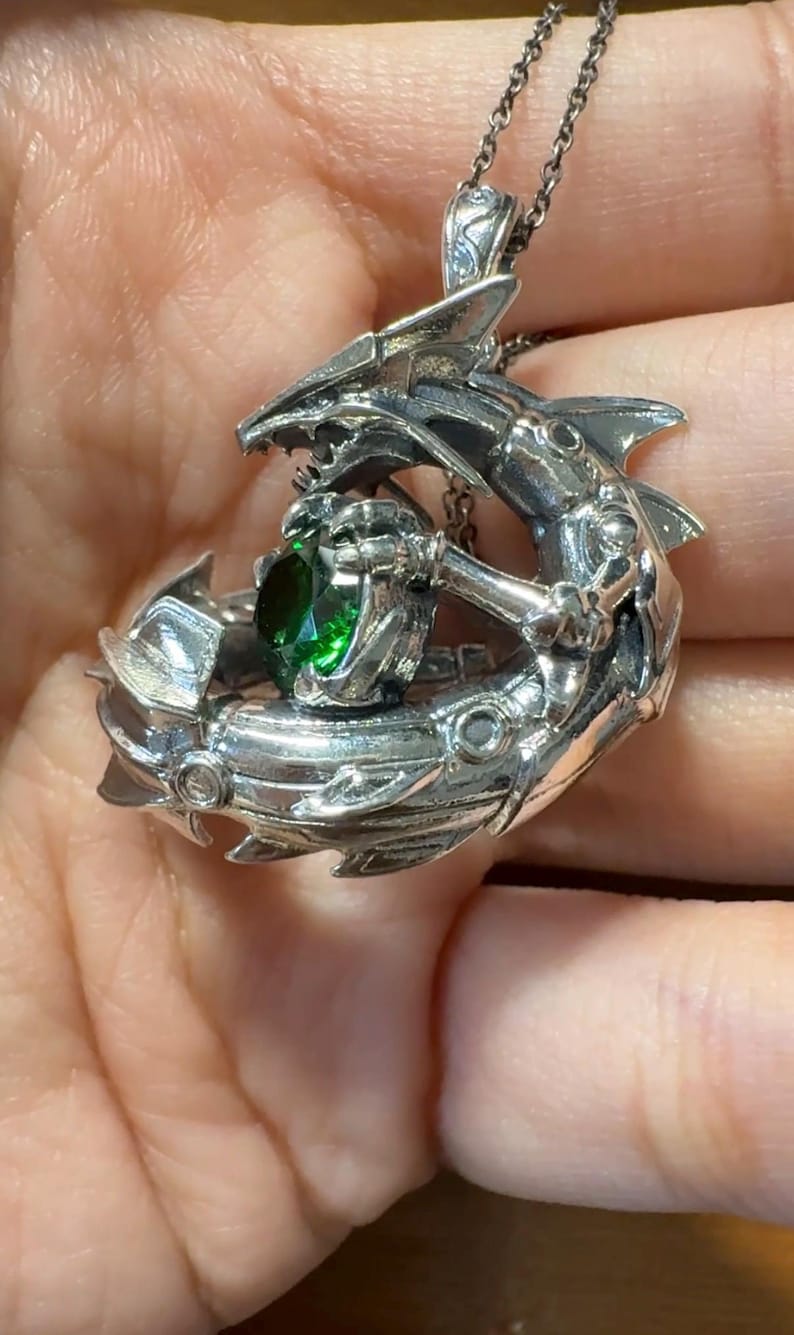 May include: A silver pendant necklace featuring a detailed, stylized shark design. The shark holds a large, round, emerald-green gemstone. The pendant hangs from a delicate silver chain, showcasing intricate metalwork and a unique, aquatic theme.