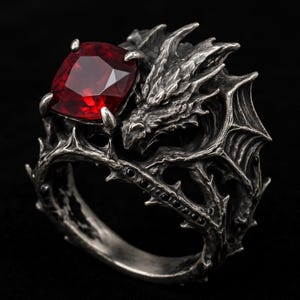 以下が含まれることがあります： 銀色のリングで、ドラゴンのデザインが特徴です。 大きな正方形の赤い宝石があり、ドラゴンの鱗と黒いアクセントで詳細に描かれています。 ドラゴンの頭はリングの側面に彫刻されています。
