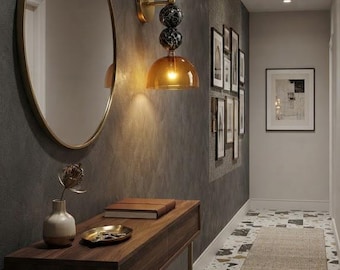 May include: A dark wood console table with thin gold legs, topped with a small vase, a gold tray, and a book. Above it hangs a round gold-framed mirror and a wall sconce featuring three black marble spheres and a translucent amber glass dome shade.