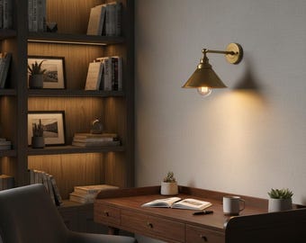 May include: A brass-colored wall-mounted lamp with a conical shade illuminates a wooden desk. The desk features a drawer, a mug, a pen, and an open book. A bookshelf filled with books and framed pictures is in the background.