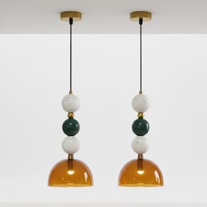Könnte beinhalten: Zwei Pendelleuchten mit modernem Design. Jede Leuchte verfügt über ein goldfarbenes Baldachin, ein schwarzes Kabel und eine Reihe von Marmor- und grünen Steinkugeln. Der Boden jeder Leuchte hat einen warmen, bernsteinfarbenen Glasschirm.